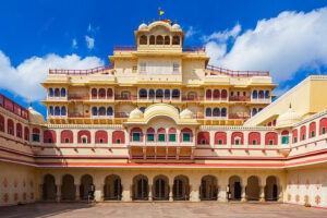 “Facade of Chandra Mahal in Jaipur’s City Palace: a grand cream-colored multi-storey palace blending Rajput and Mughal architectural features—arched galleries, jharokha balconies, and ornate detailing—under a bright blue sky